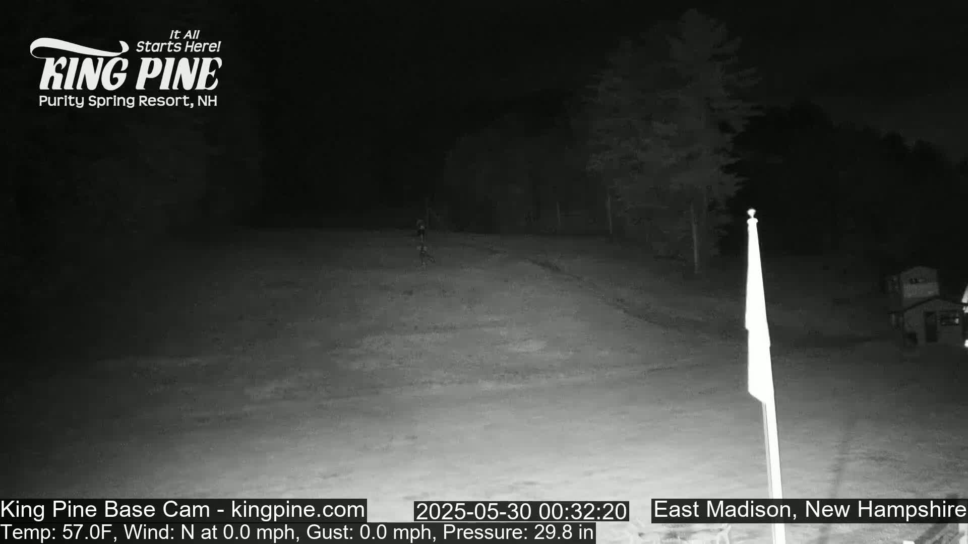 A nighttime, low-light view of a mostly empty snow-covered field with trees and a flagpole in the background under a clear sky.