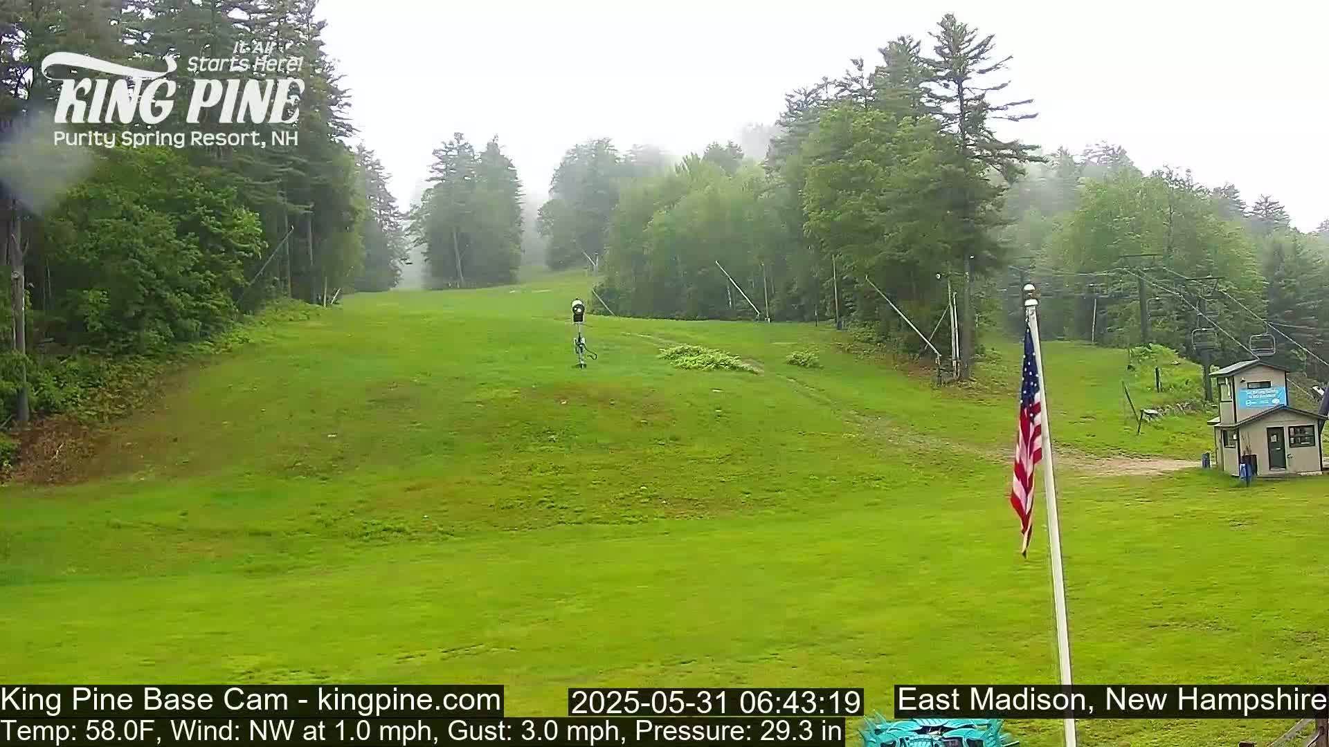 A grassy ski slope is visible under an overcast sky, with a small building and an American flag in the foreground.