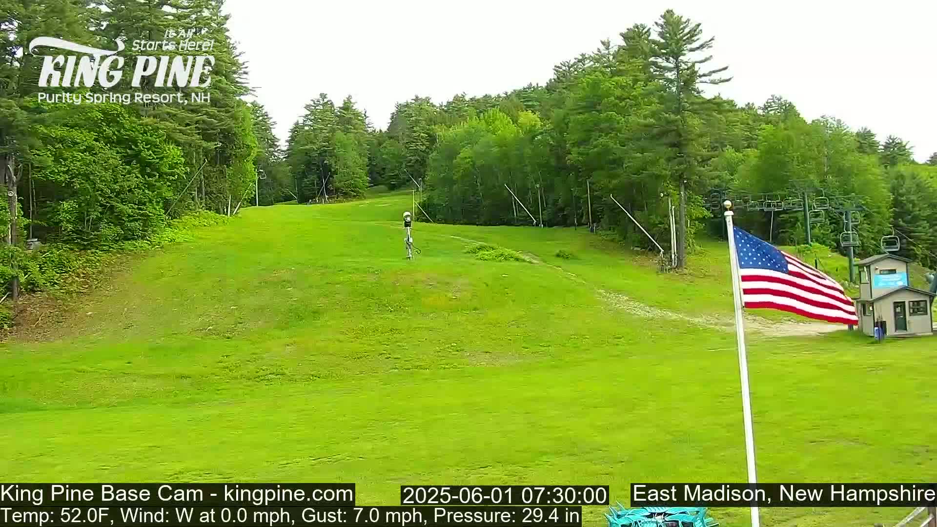A grassy ski slope with a chairlift and an American flag is visible on a partly cloudy day.