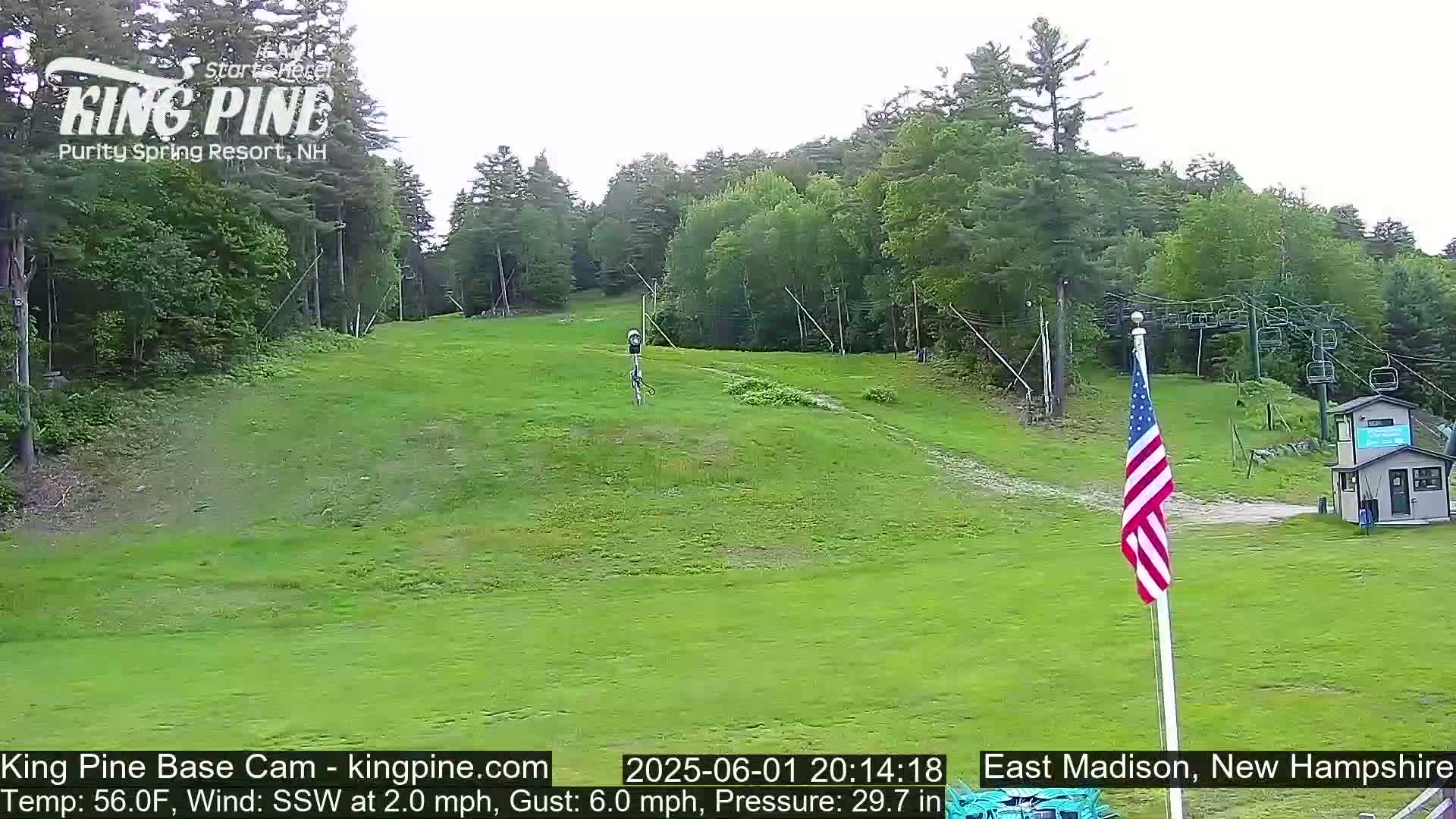 A grassy ski slope, mostly empty, with a chairlift and an American flag visible under a partly cloudy sky.