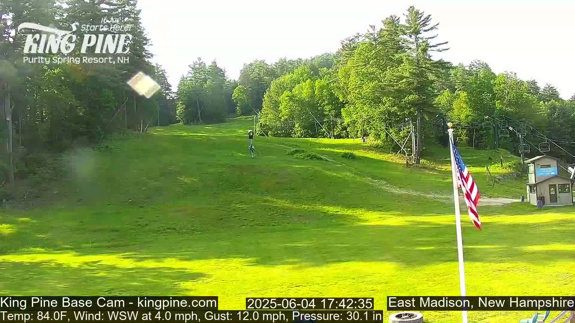 A grassy ski slope, surrounded by green trees under a mostly sunny sky, features a chairlift, a small building, and an American flag.