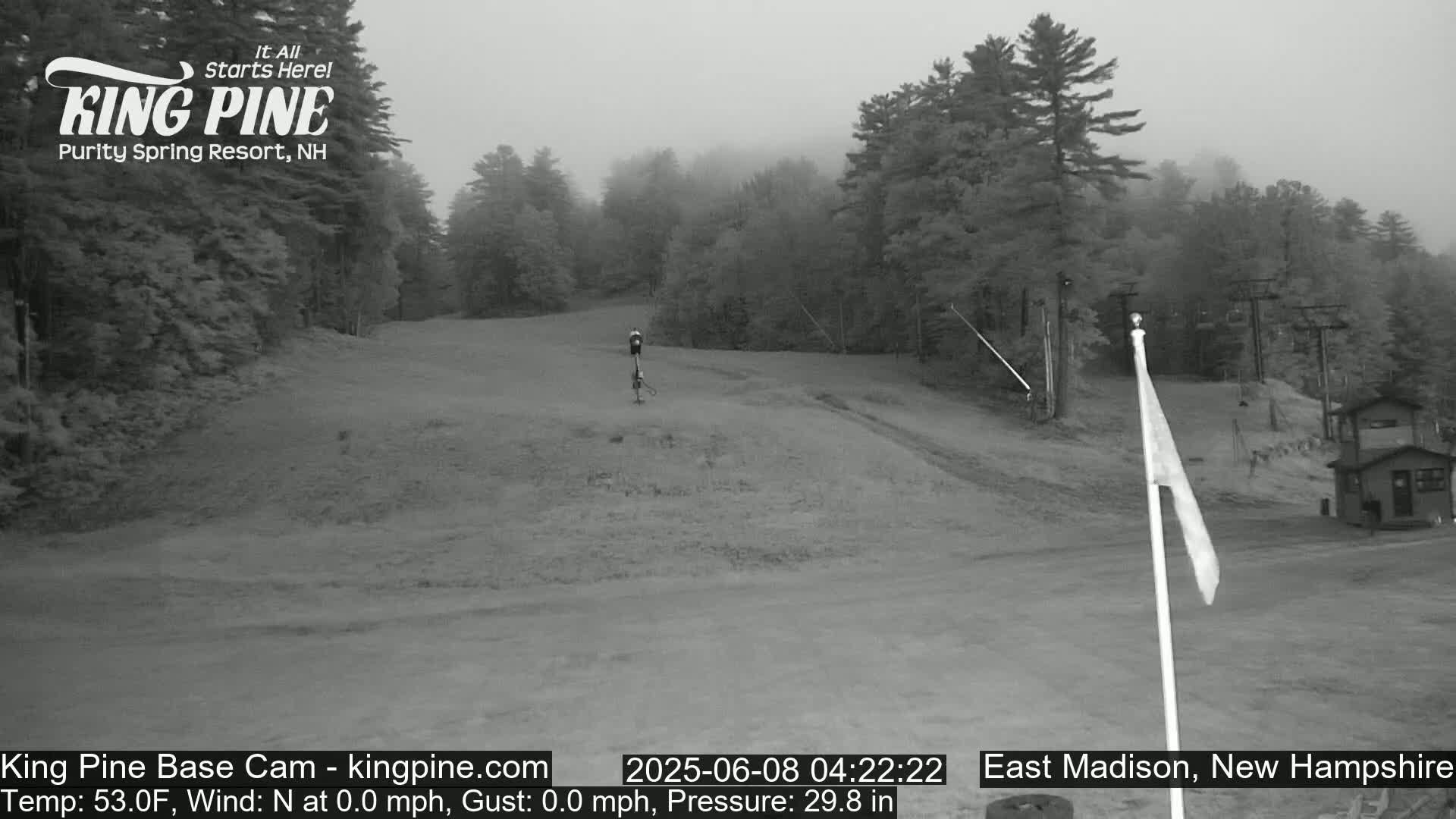 A foggy, grayscale view of a ski slope with a chairlift and a small building in the background.