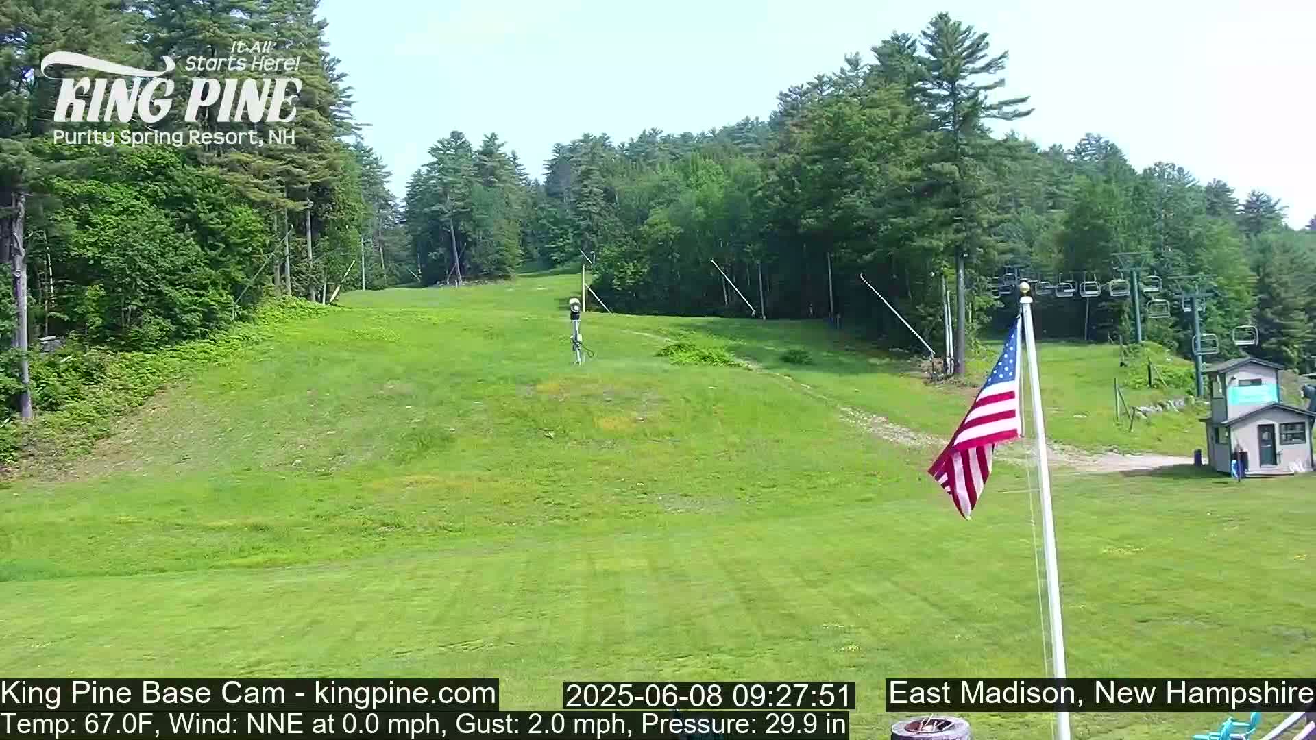 A grassy ski slope with a chairlift and American flag, surrounded by green trees under a clear sky.