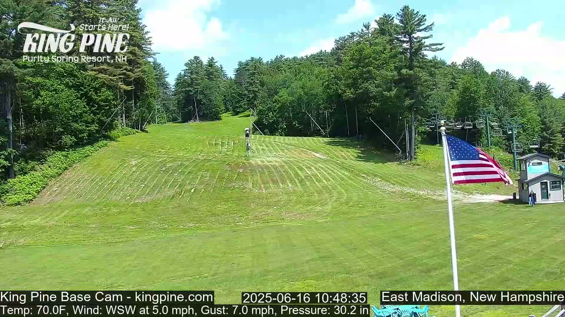 A grassy ski slope, mostly mowed, with a chairlift and an American flag visible in the background on a partly sunny day.