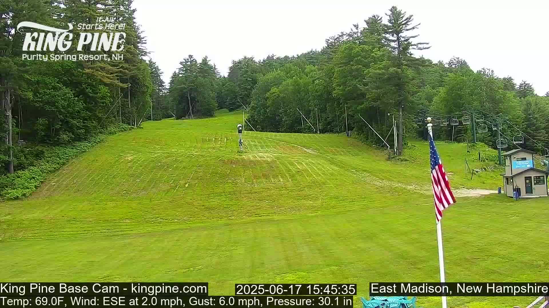 A grassy ski slope with a chairlift and American flag in the background on a partly cloudy day.
