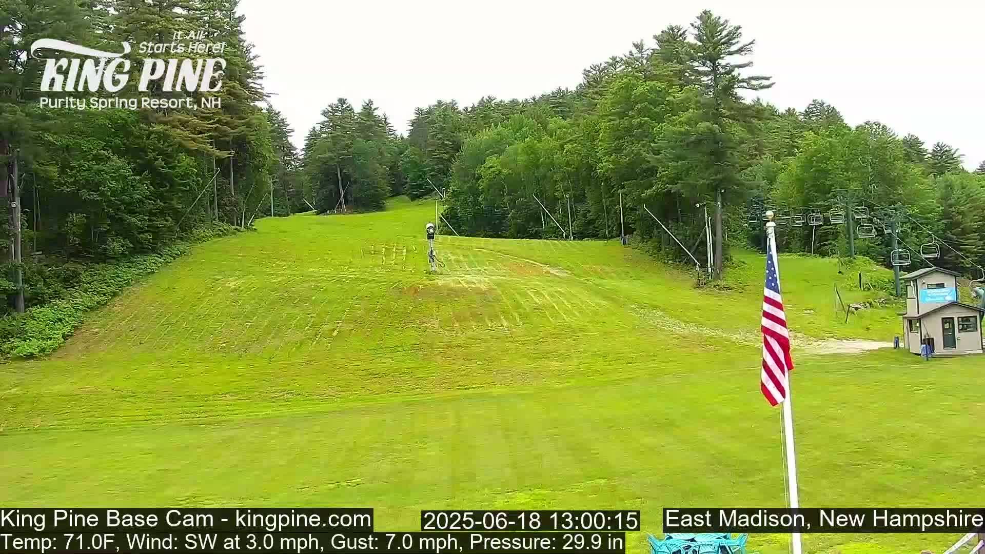 A grassy ski slope is visible, with a chairlift and an American flag in the foreground, under partly cloudy skies.