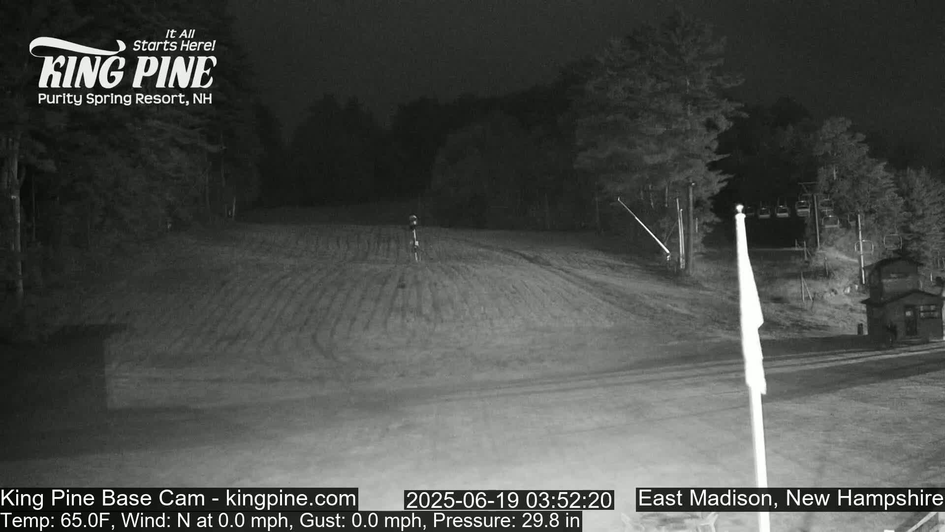 A nighttime, low-light view of a mostly empty ski slope with calm conditions, a few trees, and a flagpole in the foreground.