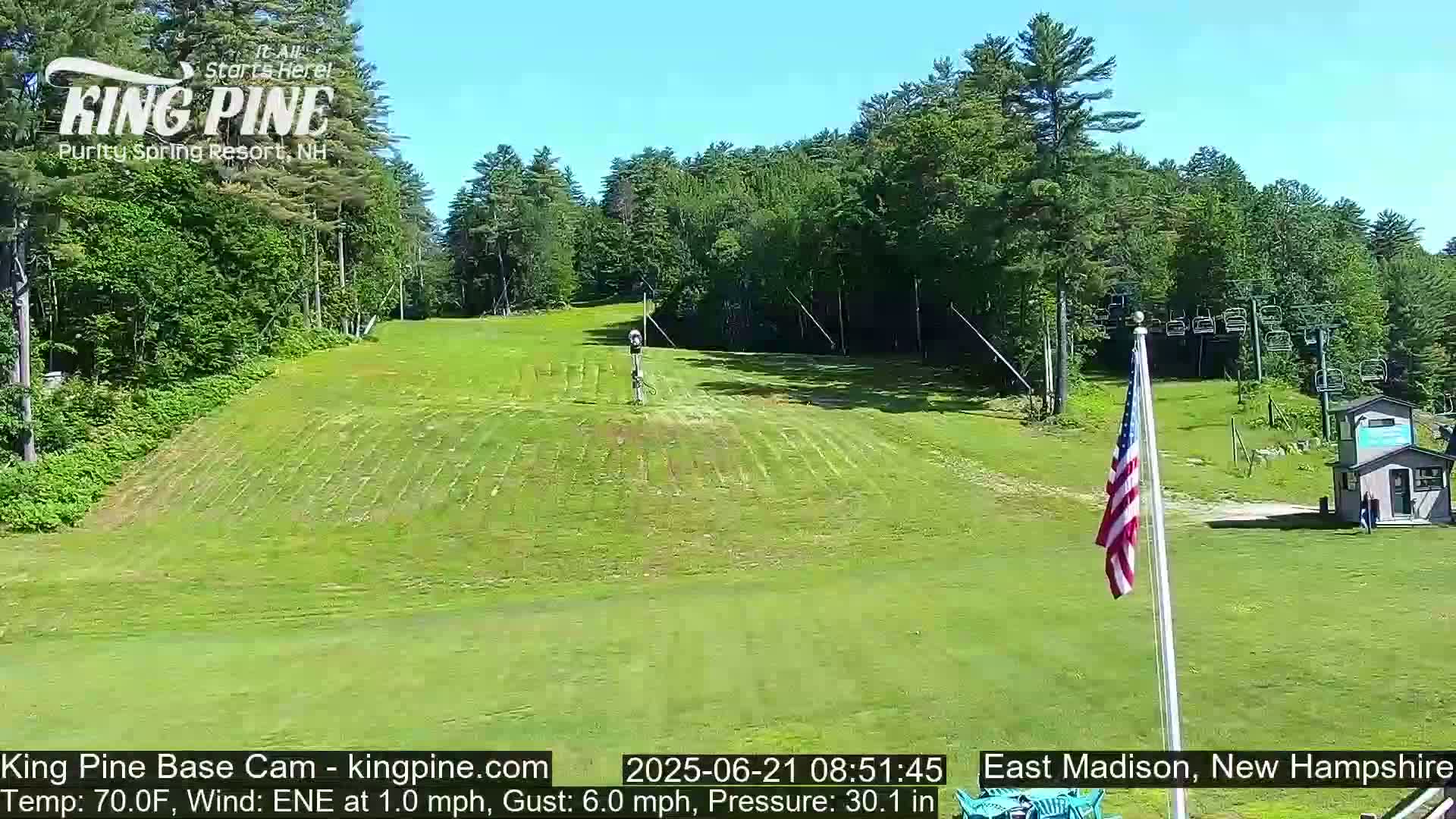 A grassy ski slope is visible under a clear blue sky, with a ski lift and an American flag in the foreground.