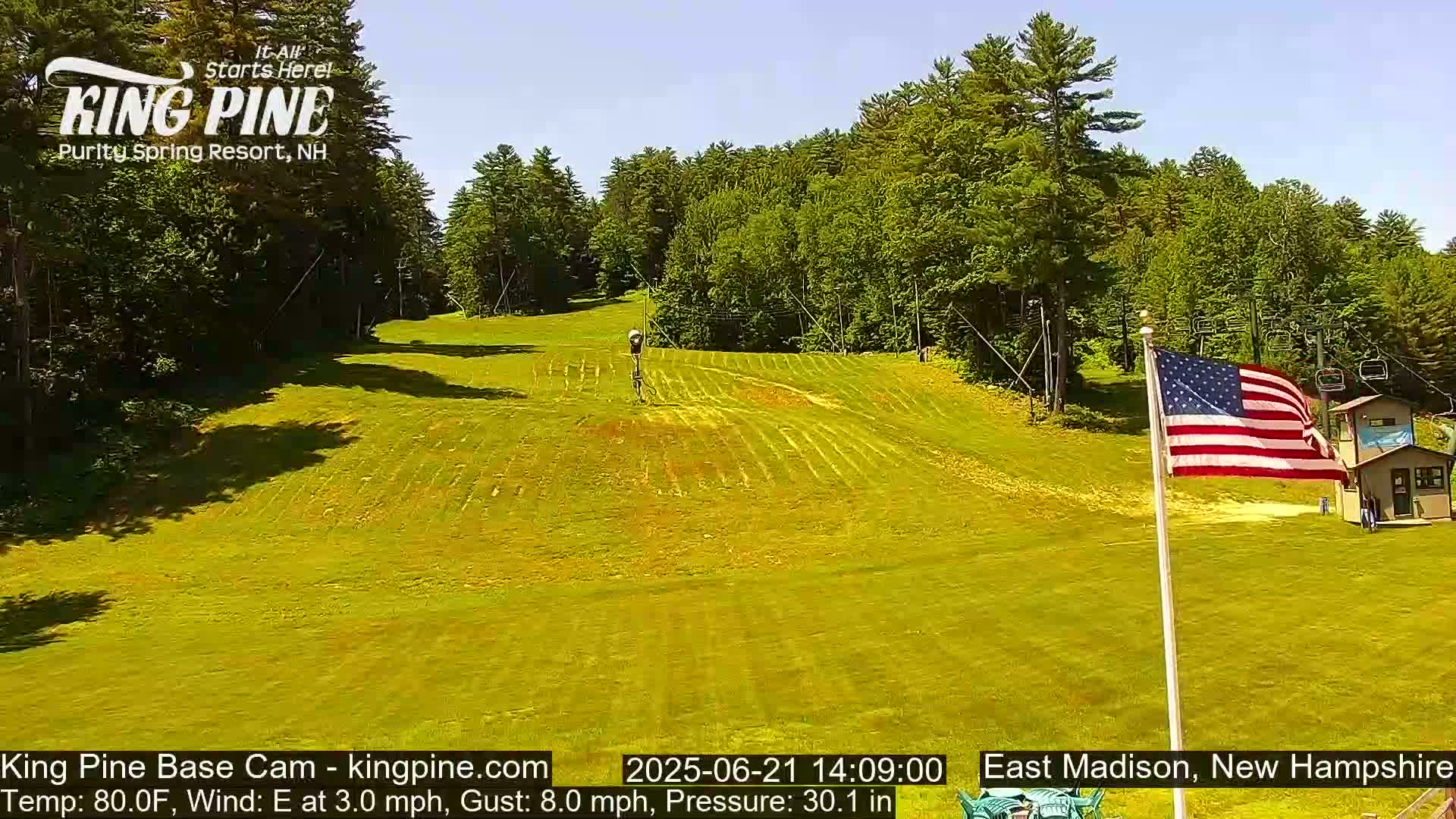 A grassy ski slope with a chairlift and an American flag waving under a clear, sunny sky.