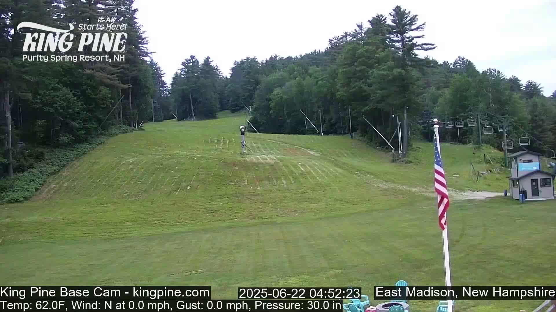A grassy ski slope is visible, with a chairlift and an American flag in the foreground, under a mostly clear sky.
