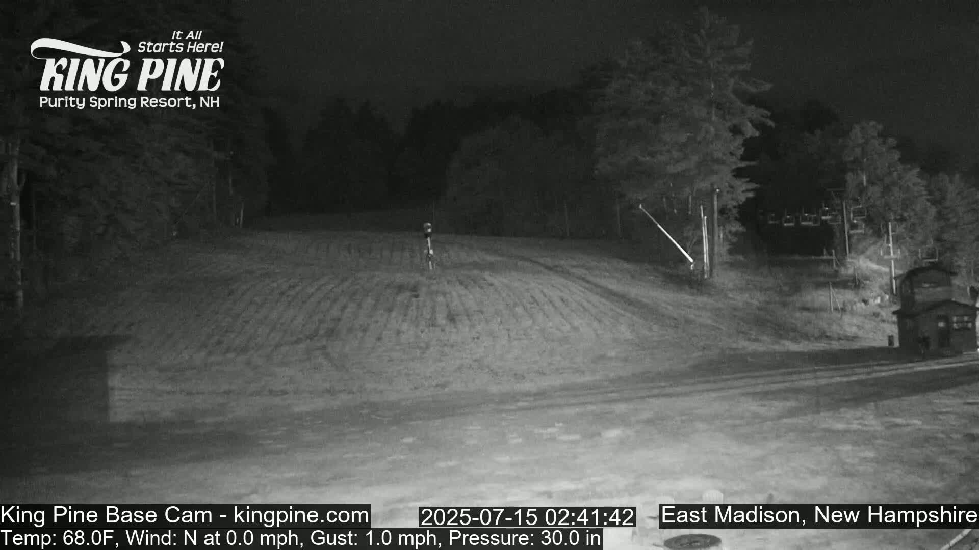 A nighttime, low-light view of a gently sloping, grassy area with trees in the background and a small building to the right, under calm conditions.
