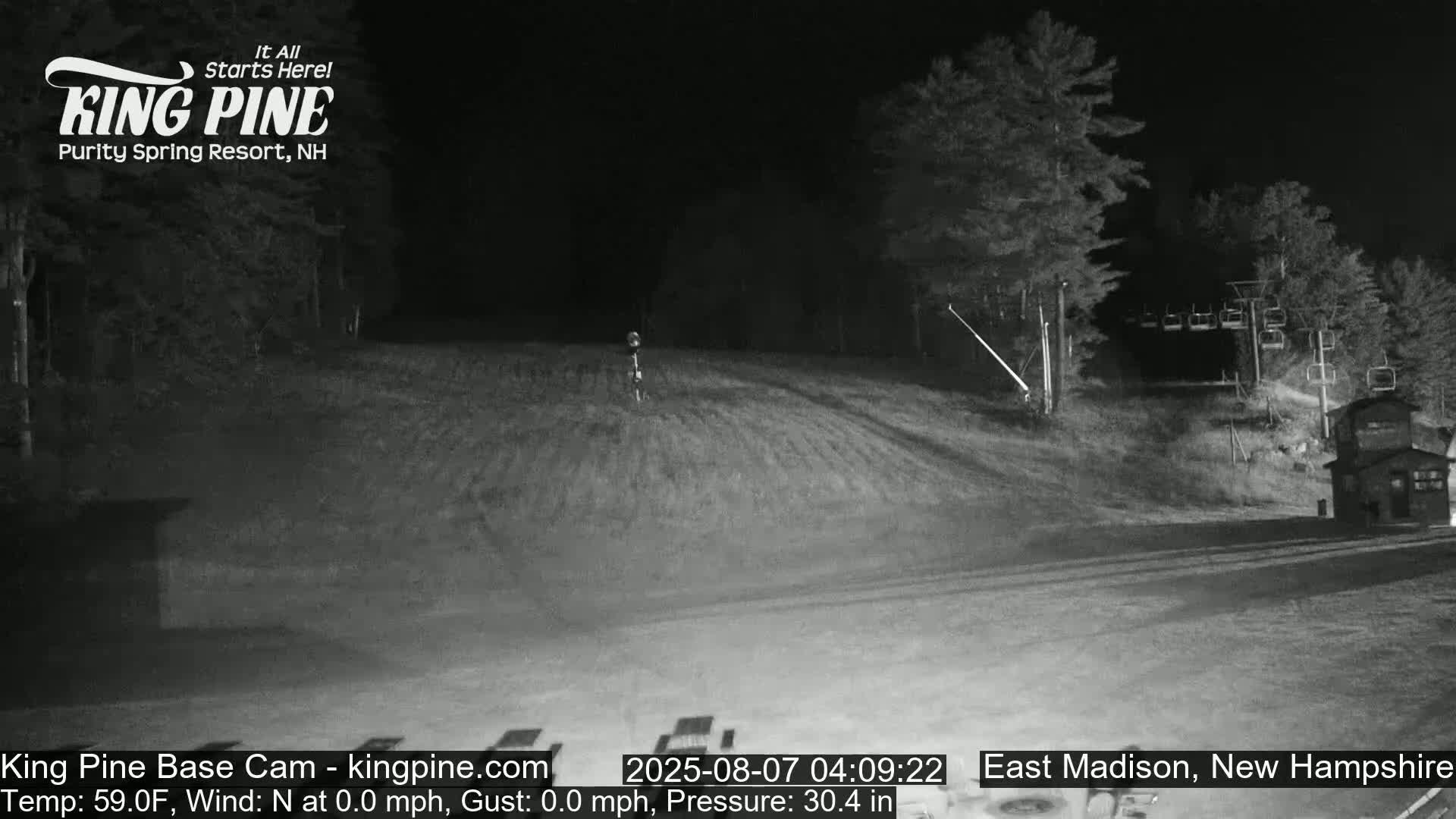 A nighttime, low-light view of a ski resort hill with a chairlift, trees, and a small building under a calm, clear sky.