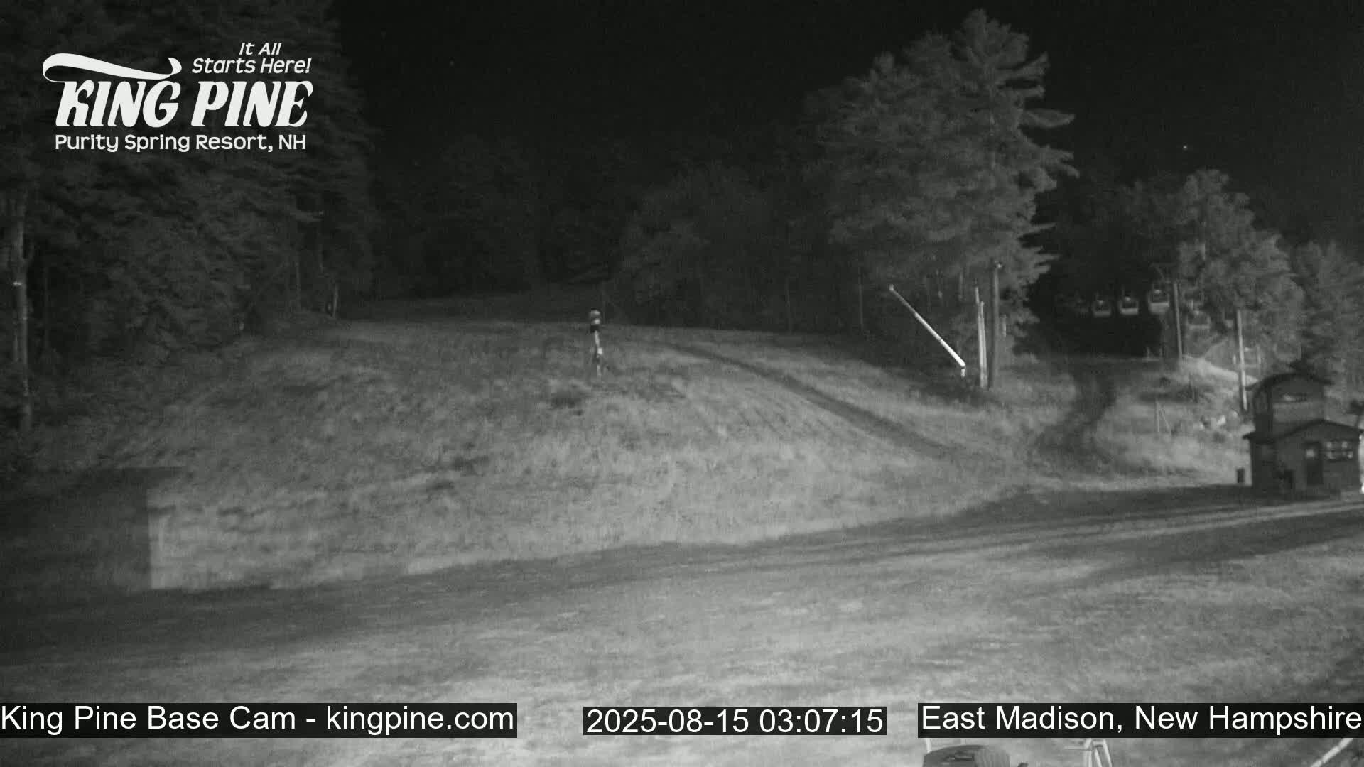 A nighttime, low-light view of a grassy hill leading up to a dark wooded area with a small building and a chairlift visible in the distance.