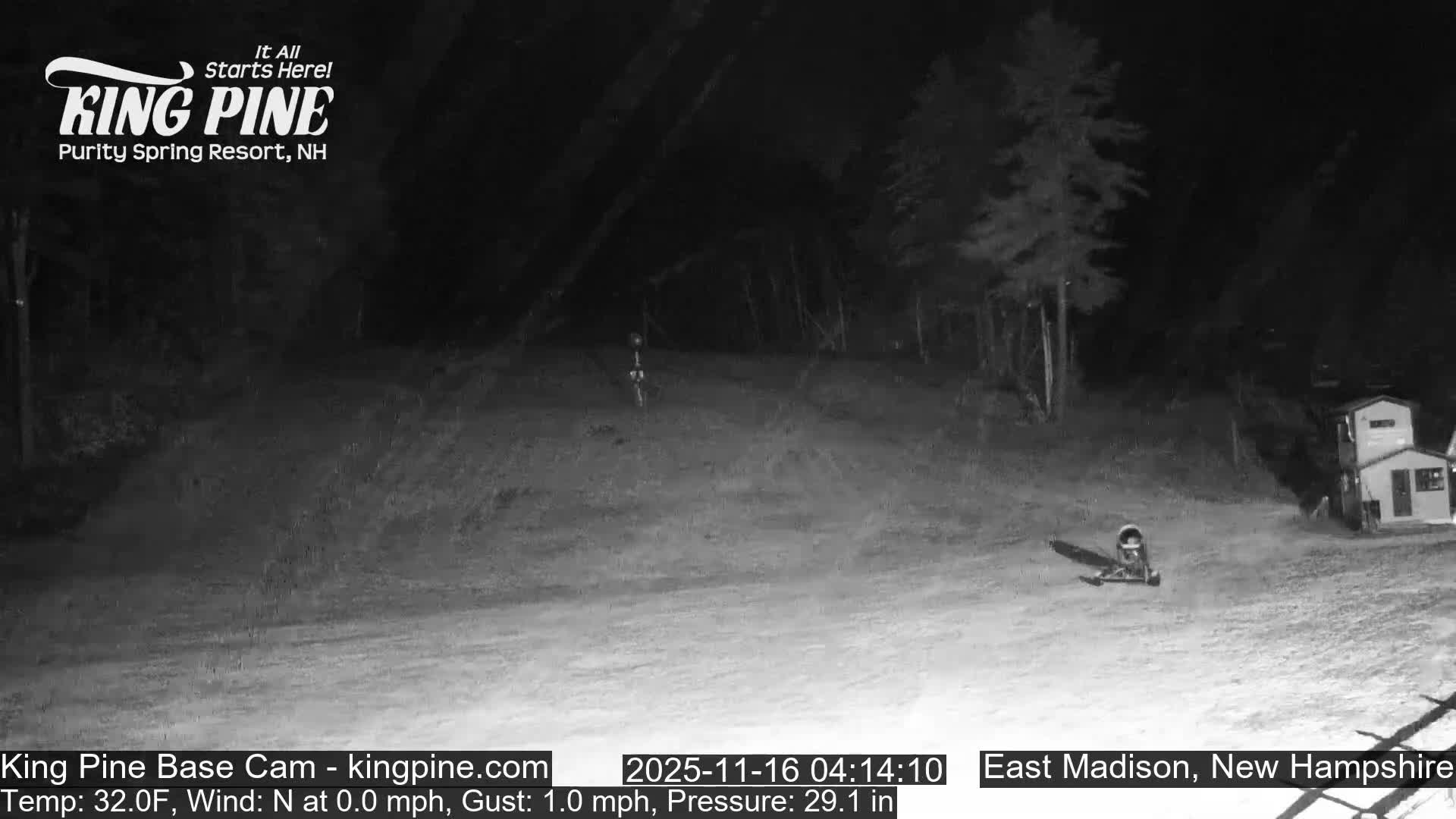 A clear, dark night is depicted in a grayscale outdoor scene, showing a cleared ski slope base with distant trees, a small building, and some equipment on the ground.