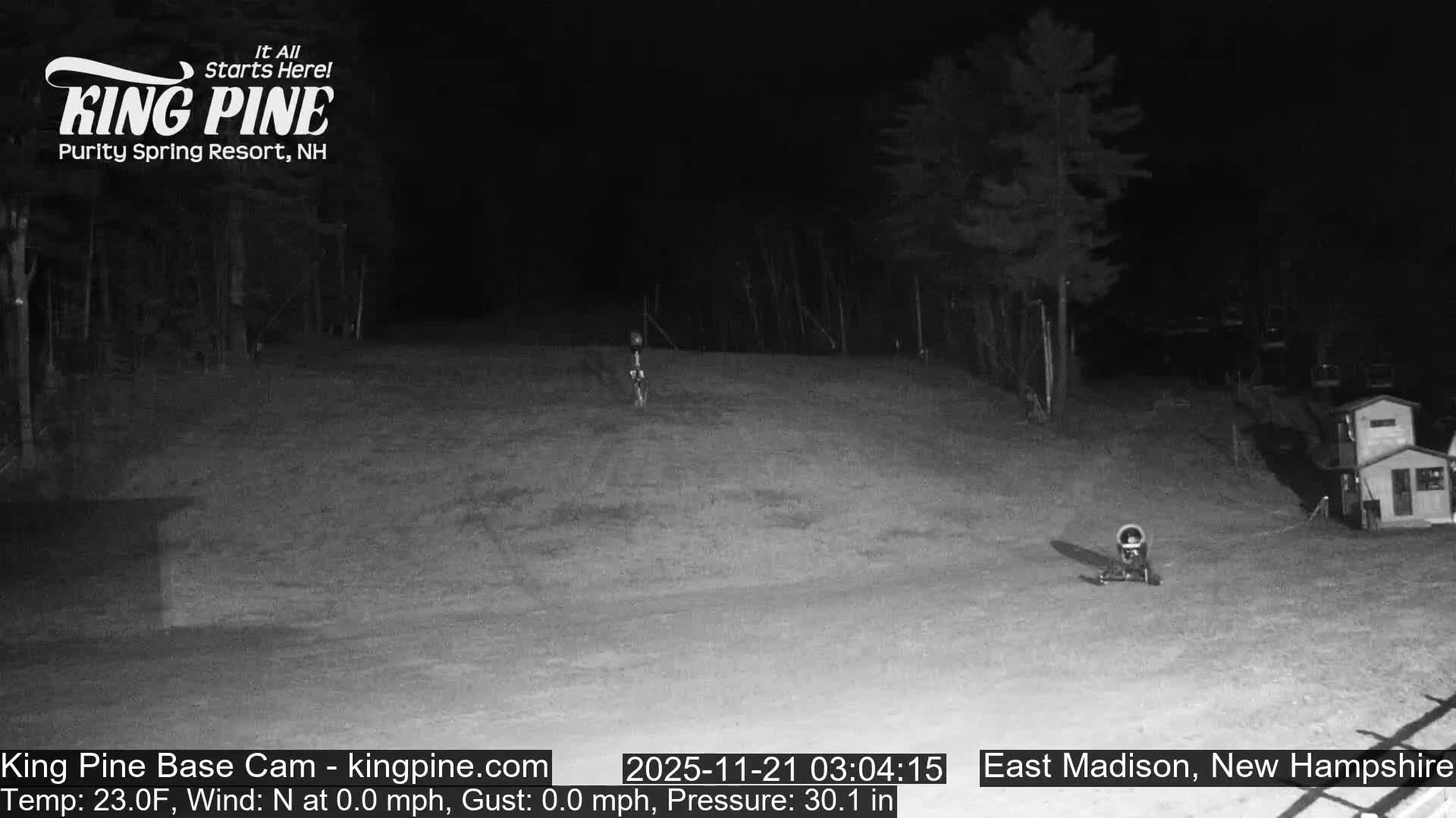 On a clear, still night with a temperature of 23.0F and no wind, a dimly lit ski slope at a resort base is visible, featuring a snow cannon, a small building, and ski lift structures on the right, all bordered by dark trees and an absence of snow.