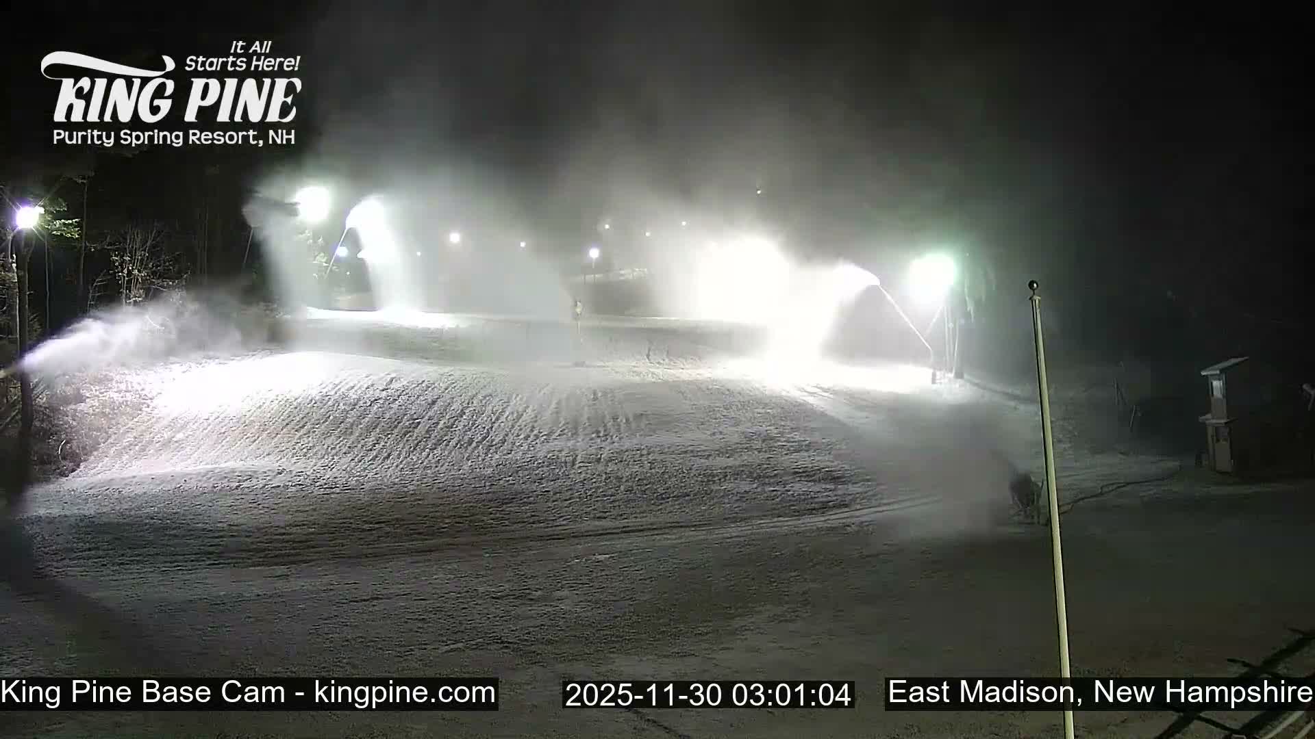 A snow-covered ski slope is brightly illuminated by artificial lights at night, with multiple snow cannons actively blowing fresh snow under clear, cold conditions.