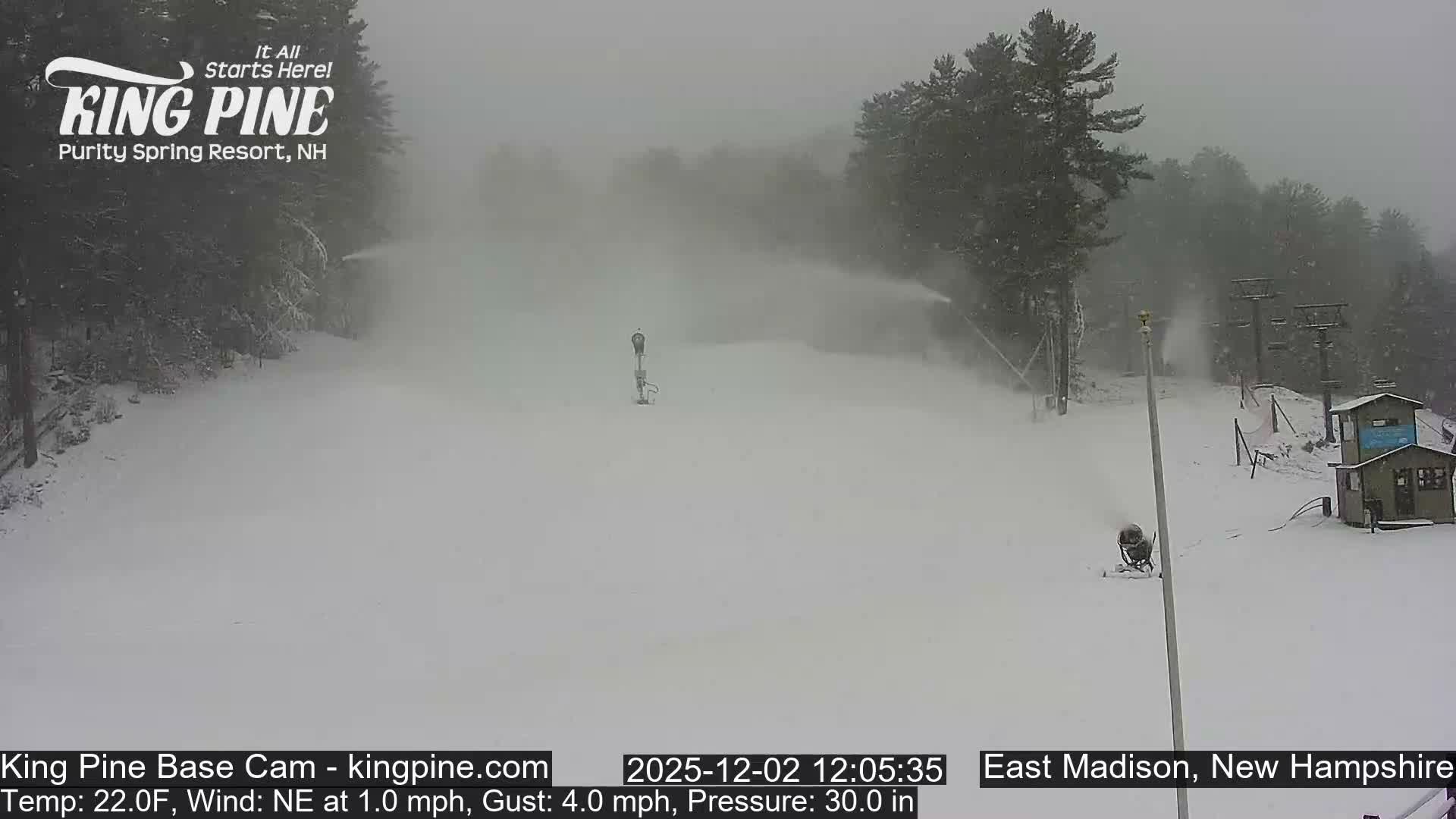 A snowy ski slope is seen under overcast and hazy conditions, with multiple snow cannons actively blowing snow, along with ski lift towers, small buildings, and snow-dusted evergreen trees.