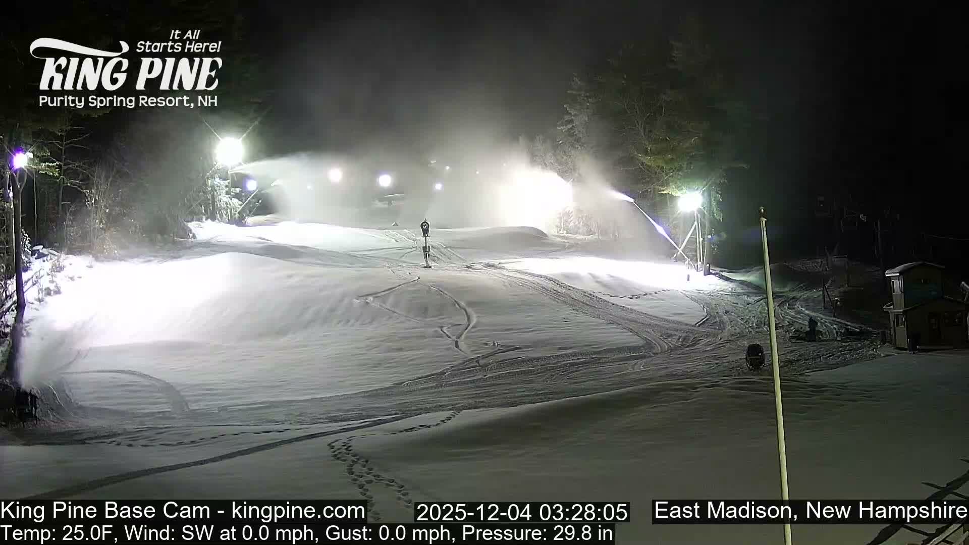 A brightly lit ski slope at night is covered in fresh snow being made by multiple active snow guns, with ski tracks and footprints visible, under calm conditions and a temperature of 25.0F.