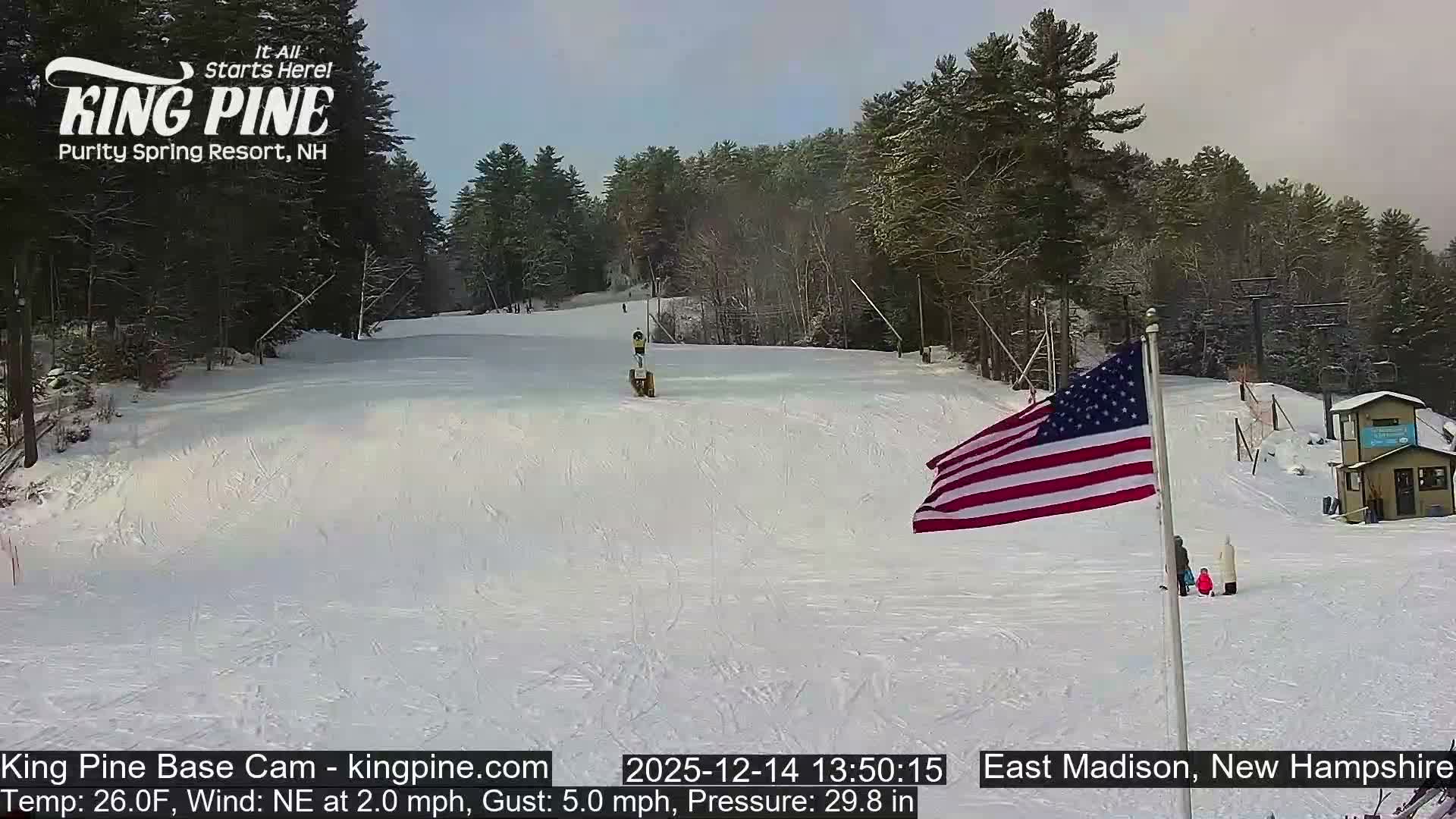 A brightly lit ski slope at night is covered in fresh snow being made by multiple active snow guns, with ski tracks and footprints visible, under calm conditions and a temperature of 25.0F.