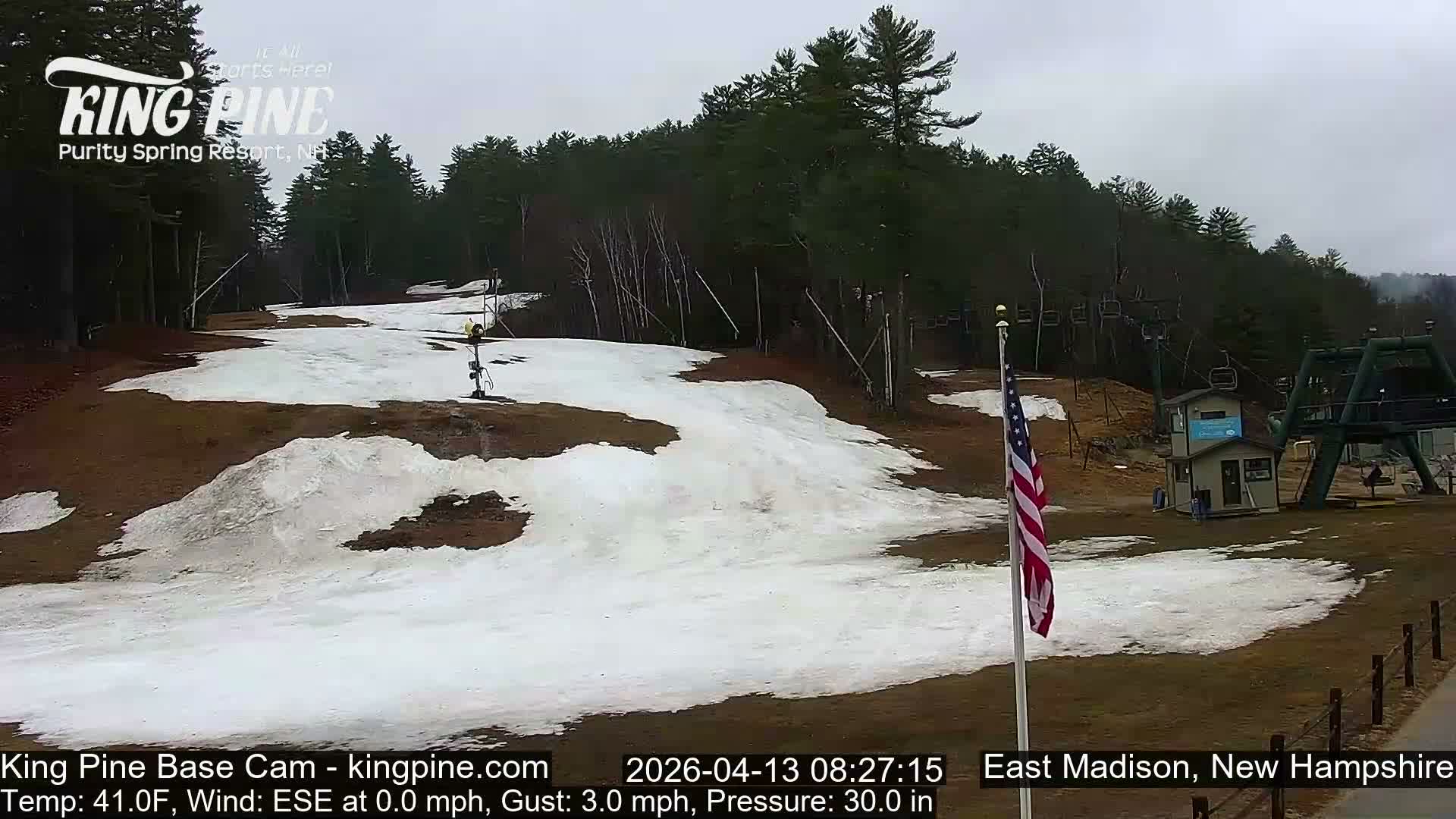 A brightly lit ski slope at night is covered in fresh snow being made by multiple active snow guns, with ski tracks and footprints visible, under calm conditions and a temperature of 25.0F.