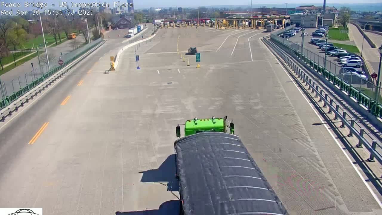 A green semi-truck approaches a mostly empty US border inspection plaza on a sunny day.