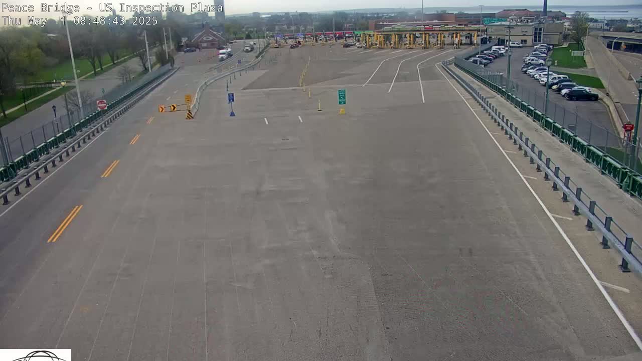 An empty asphalt roadway leads to a border crossing plaza with a few parked cars under a partly cloudy sky.