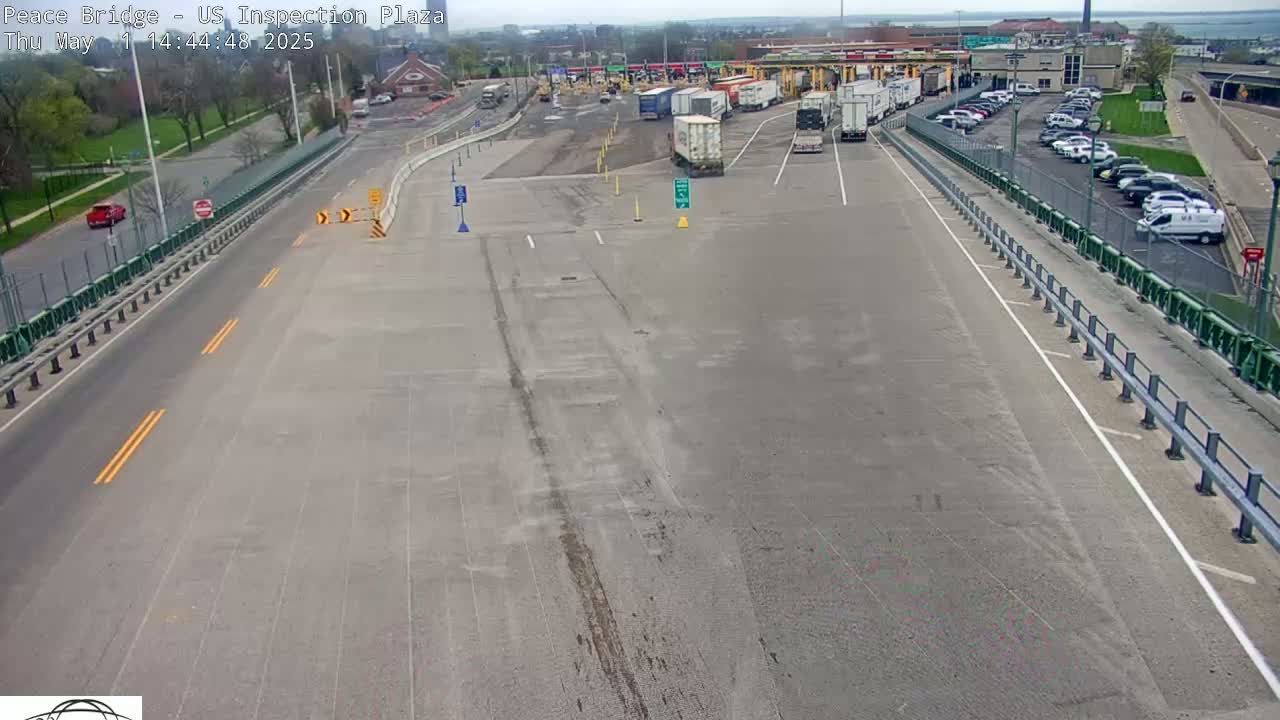 A mostly empty highway approaches a border inspection plaza on a partly sunny day, with several vehicles parked in the plaza's lot and a few more driving on the road.