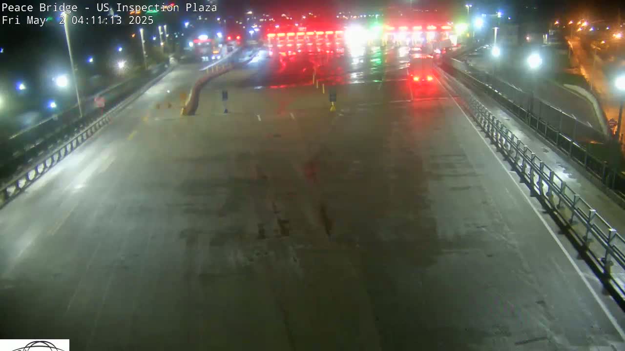A wet, empty roadway at night leads to a border crossing inspection plaza with a few vehicles visible in the distance under bright lights.