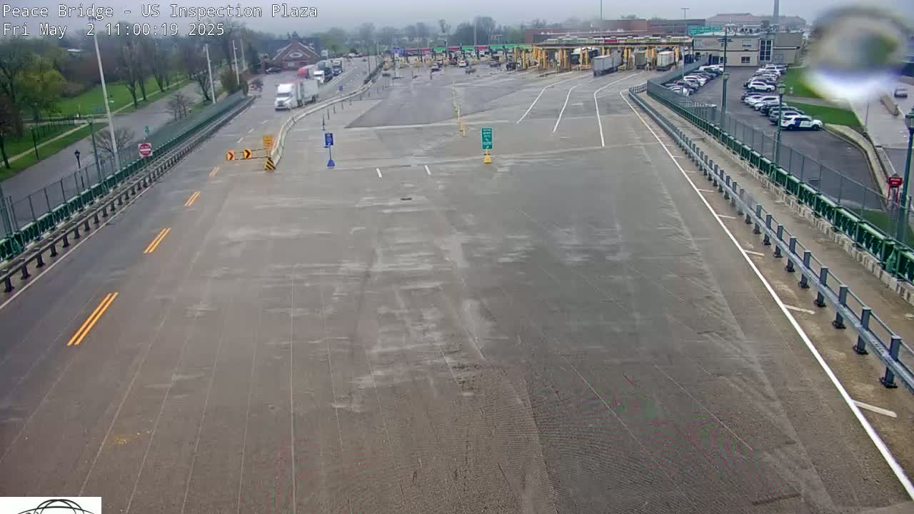 A wet roadway leads to a border inspection plaza with several vehicles parked and a few trucks driving.