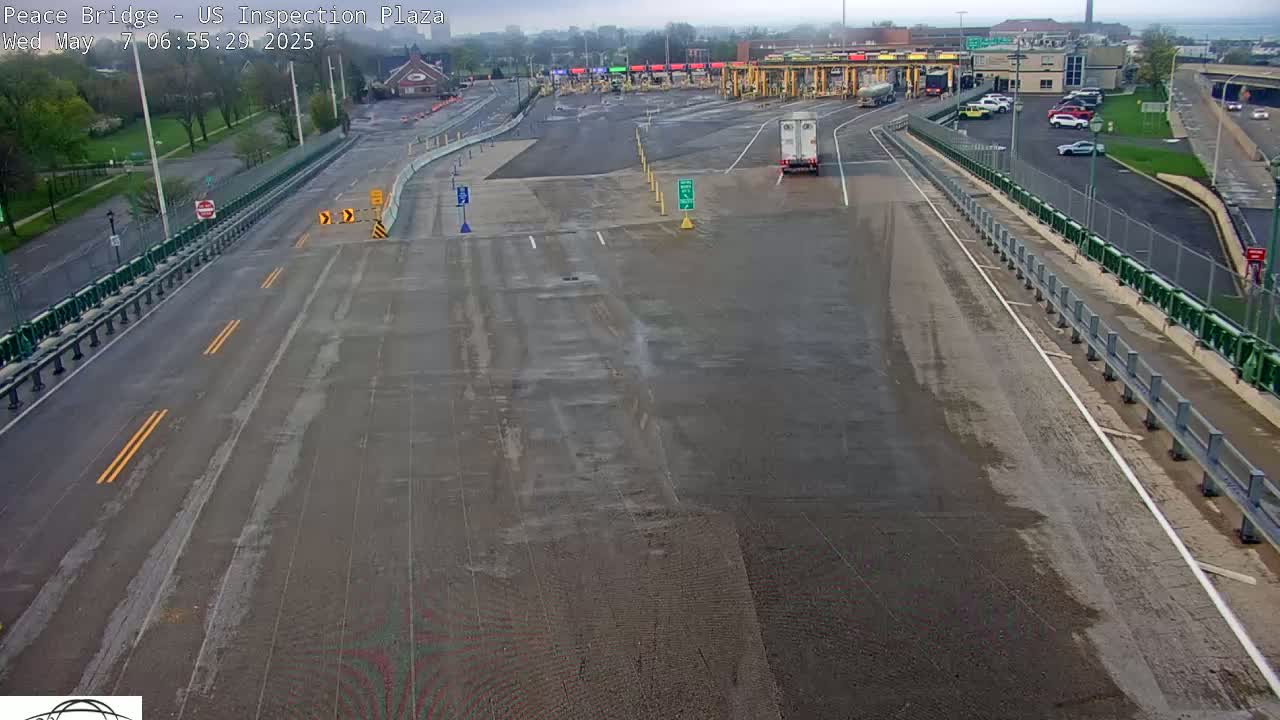 A wet, empty roadway approaches a border inspection plaza with a few vehicles visible in the distance.