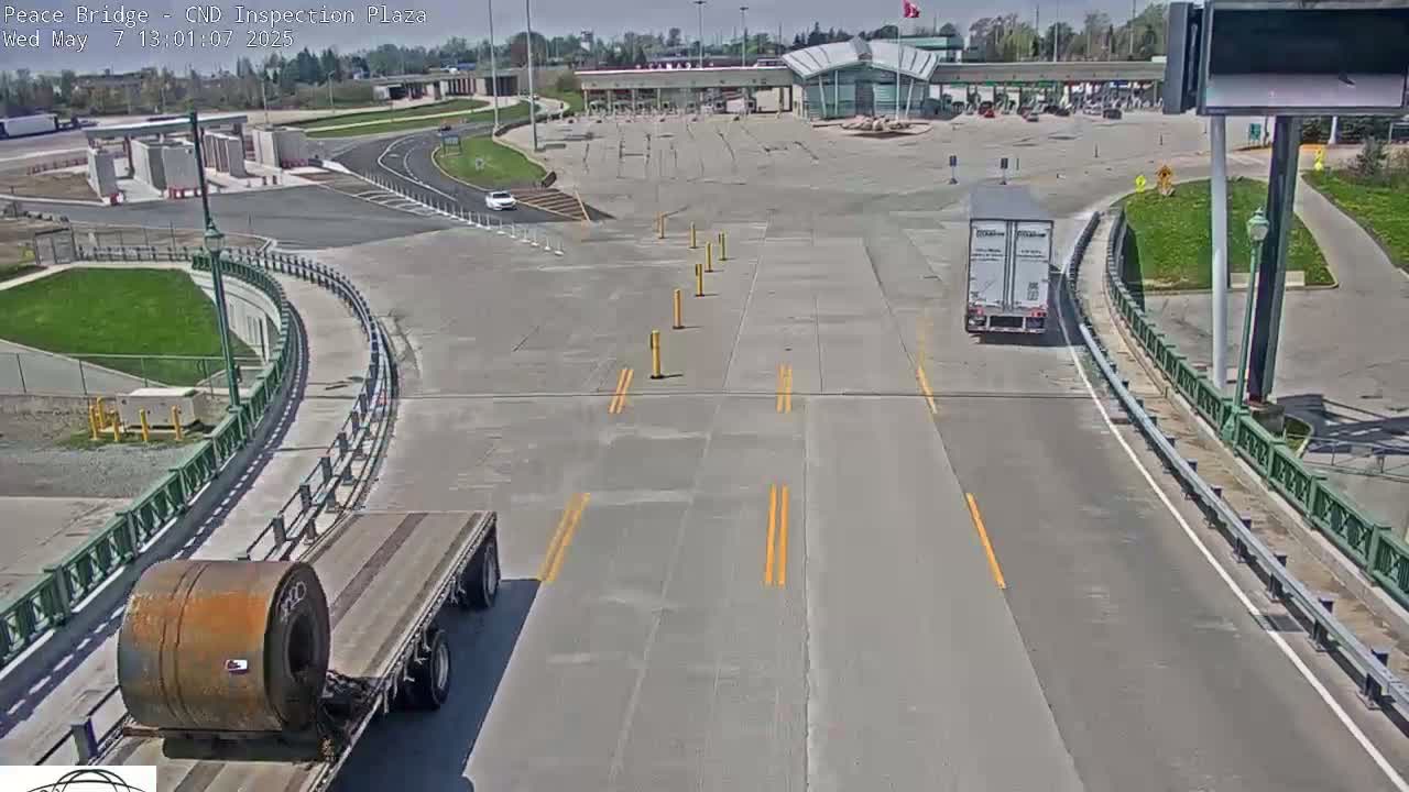 A high-angle view shows a mostly empty border crossing inspection plaza on a sunny day, with a flatbed truck carrying a large roll of metal and a semi-truck in the foreground.