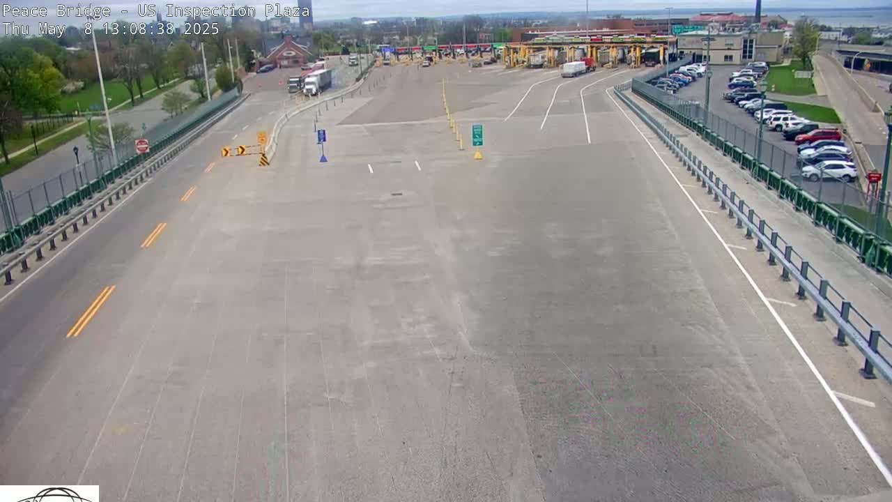 A mostly empty highway approaches a border inspection plaza on a sunny day, with several cars parked in a lot nearby.