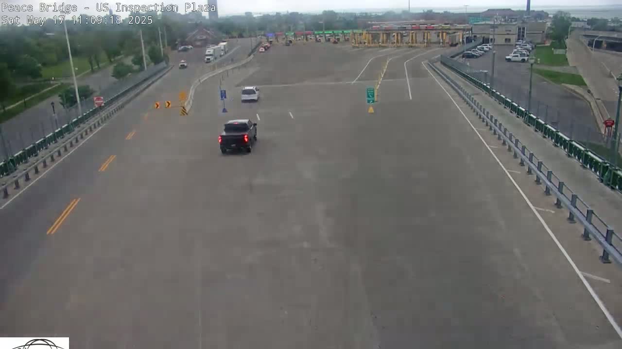 A dark-colored pickup truck and a white car drive toward a border inspection plaza on a mostly overcast day.