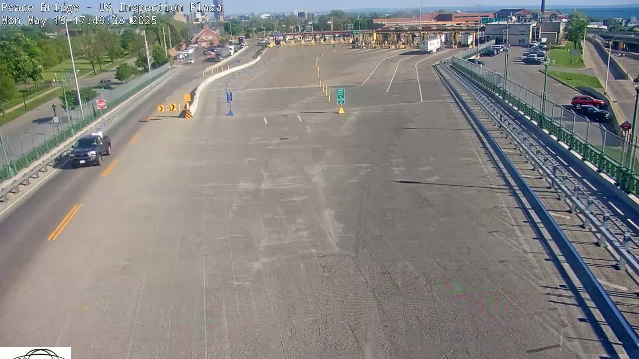 A mostly empty highway approaches a border inspection plaza on a sunny day, with a few cars visible on the road and in the parking lot.