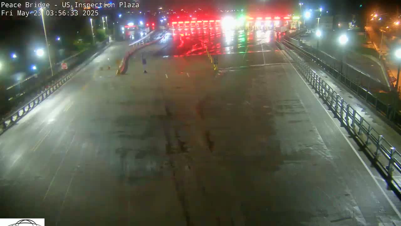A wet, empty roadway at night leads to a border inspection plaza with illuminated toll booths.