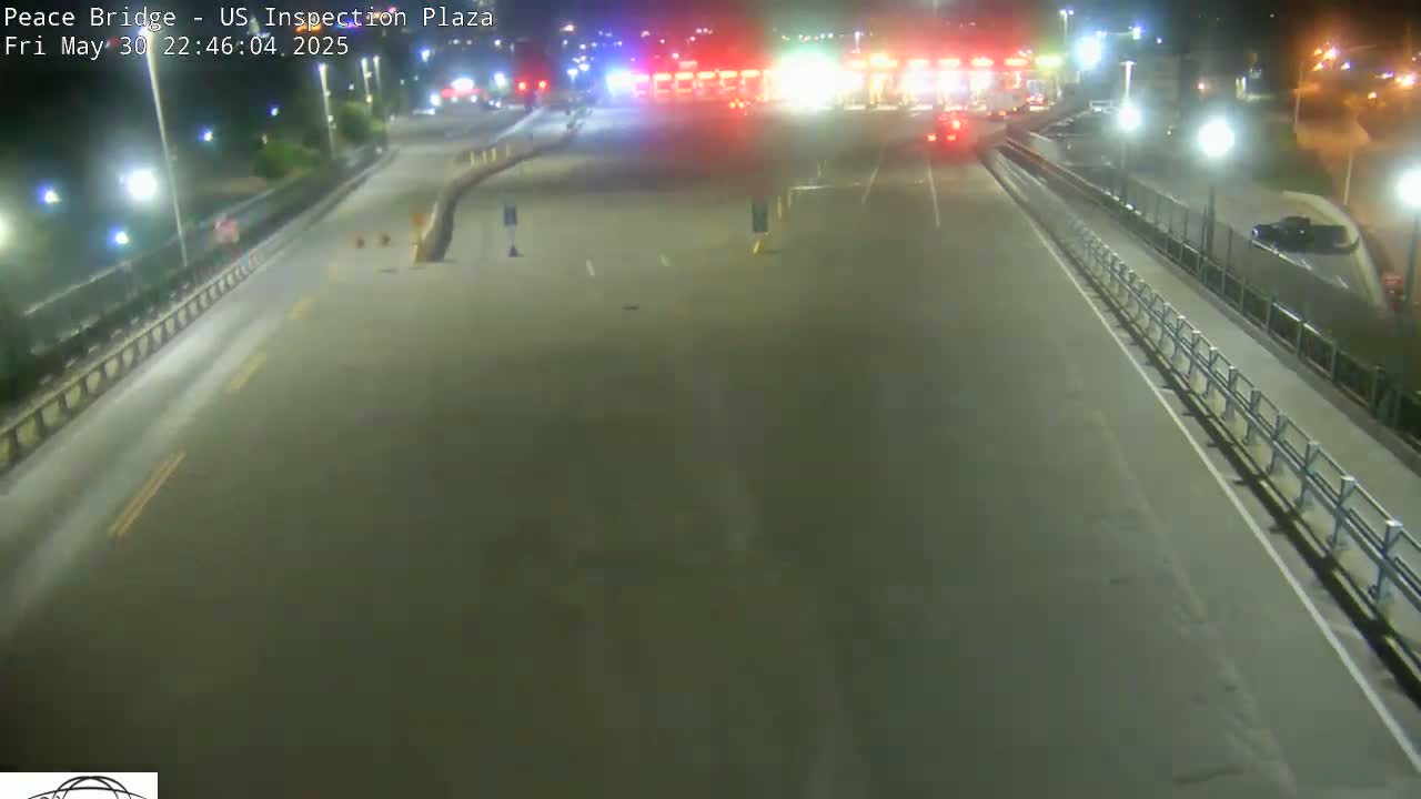 A nighttime, high-angle view shows an empty highway approaching a border inspection plaza with a few vehicles visible in the distance.