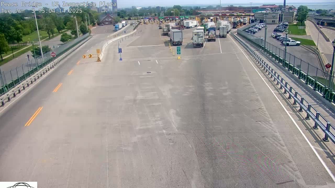A high-angle, long shot of a bridge's inspection plaza on a sunny day, showing several semi-trucks and cars in the parking area, and empty lanes leading to it.