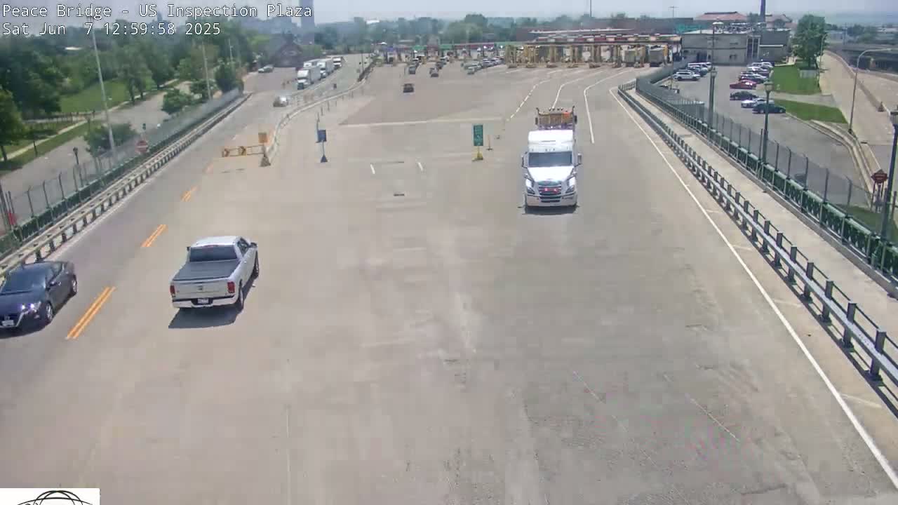 A high-angle view shows a roadway leading to a border inspection plaza on a sunny day, with several vehicles including a pickup truck and a semi-truck traveling on the road.