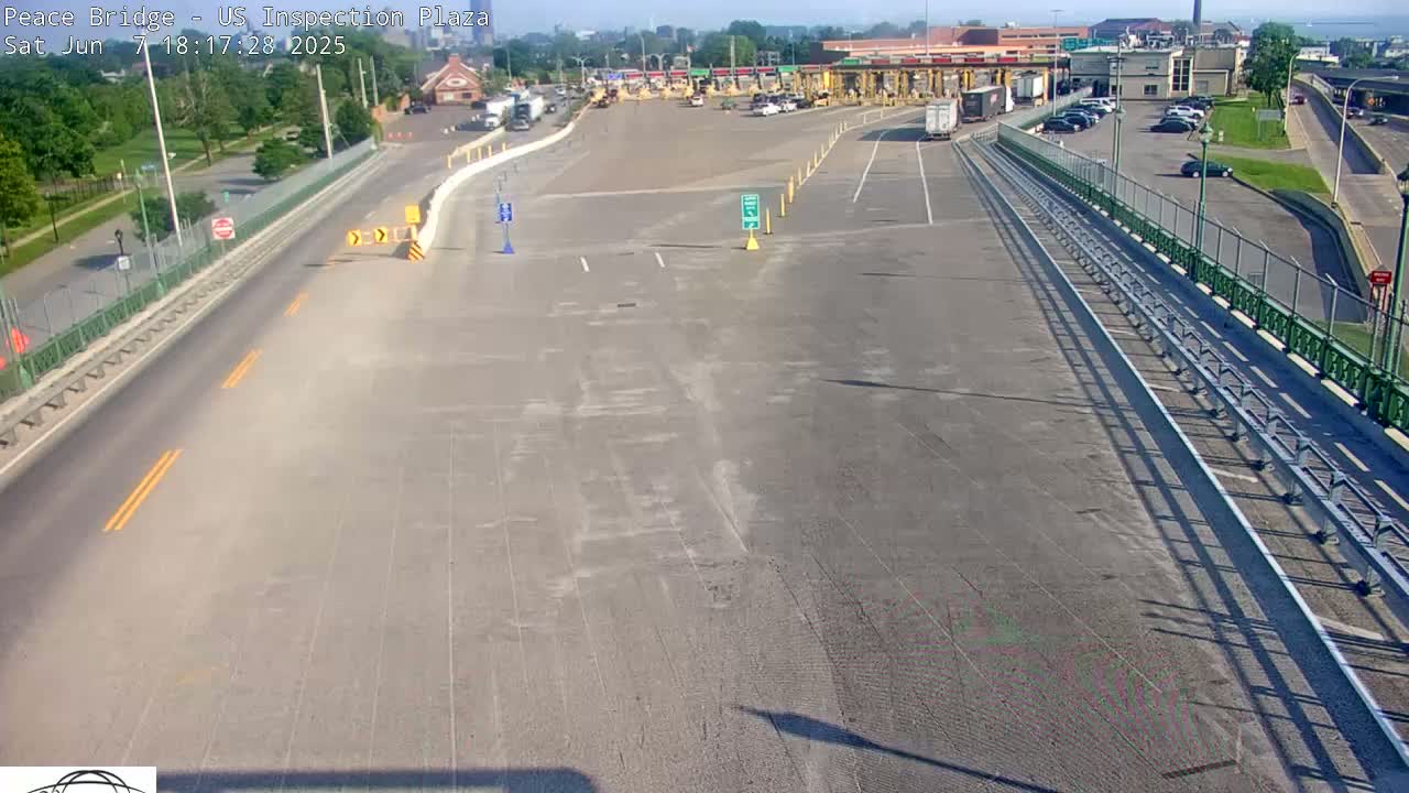 A mostly empty highway approaches a border inspection plaza on a sunny day, with a few vehicles visible in the parking lot and some greenery in the background.