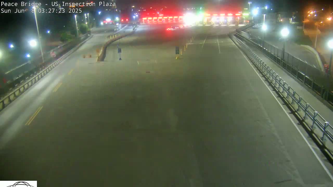 A nighttime, high-angle view shows a mostly empty roadway leading to a border inspection plaza with a few vehicles present.