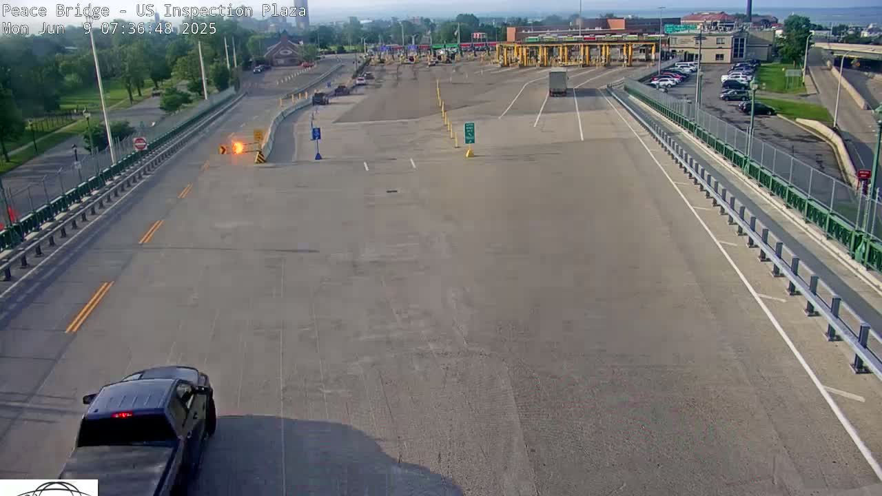 A dark-colored pickup truck approaches a mostly empty US inspection plaza on a sunny day.