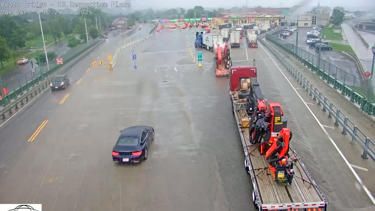 A rain-soaked roadway shows several cars and trucks, including a large flatbed carrying construction equipment, at a border crossing.