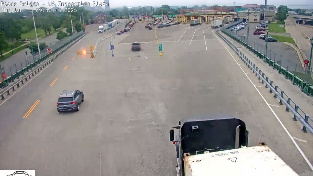 A mostly empty roadway at a border inspection plaza on a sunny day, with a few cars and trucks visible, and a small burst of fire or sparks in the middle of the road.