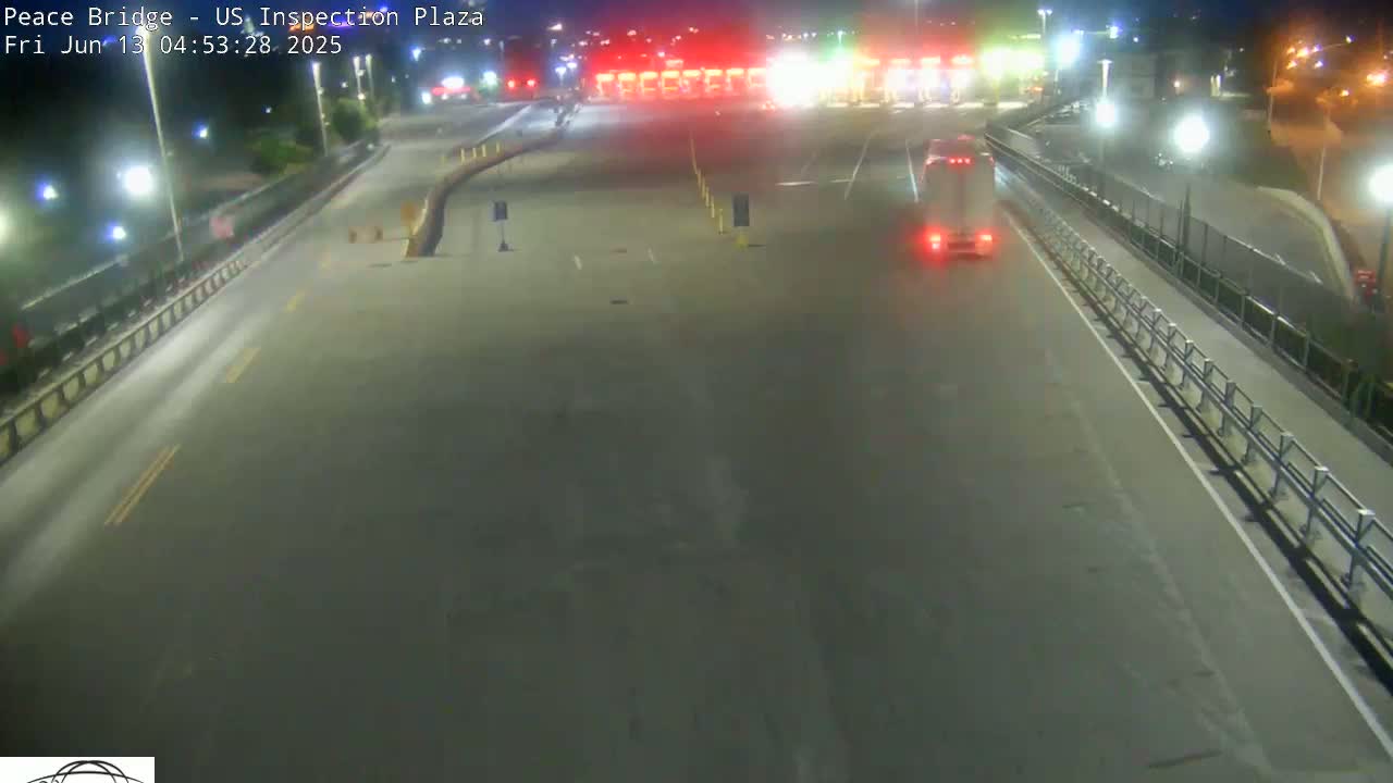 A nighttime view of a mostly empty roadway leading to a border inspection plaza, with a single vehicle approaching.