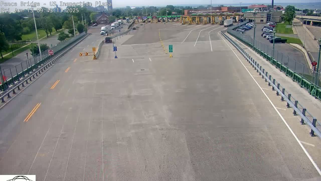 An empty highway approaches a border inspection plaza on a sunny day, with several vehicles parked in the plaza's parking lot.
