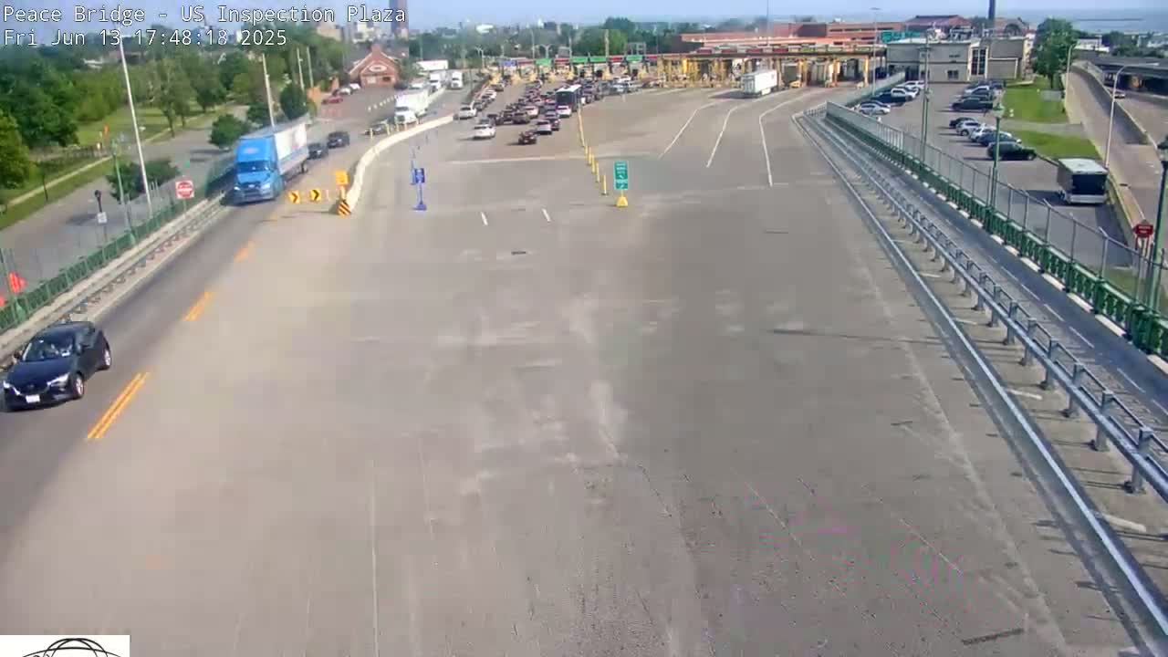 A mostly empty highway approaches a border inspection plaza on a sunny day, with several cars and trucks visible.