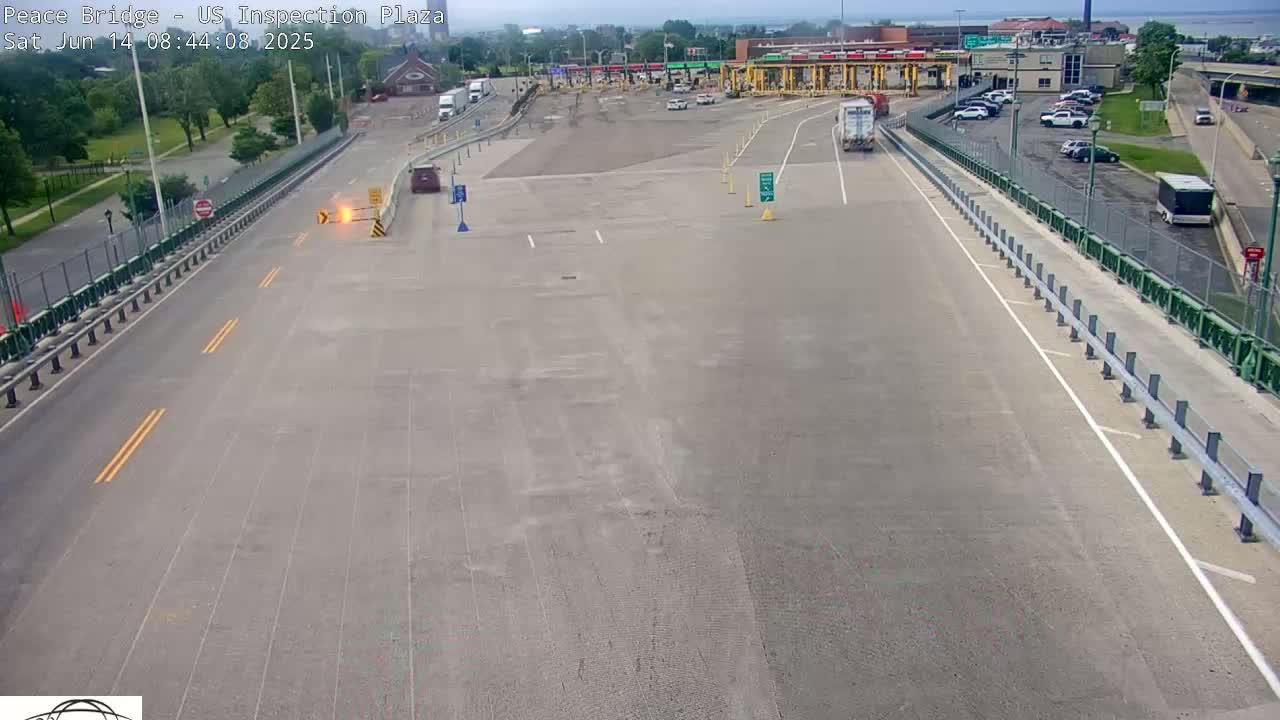 An aerial view shows a mostly empty US inspection plaza on a partly cloudy day, with several vehicles parked and a few driving on roads leading to and from the plaza.