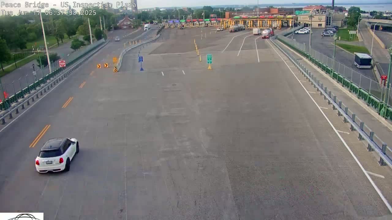 A white Mini Cooper drives on a paved road leading to a border inspection plaza on a partly sunny day.