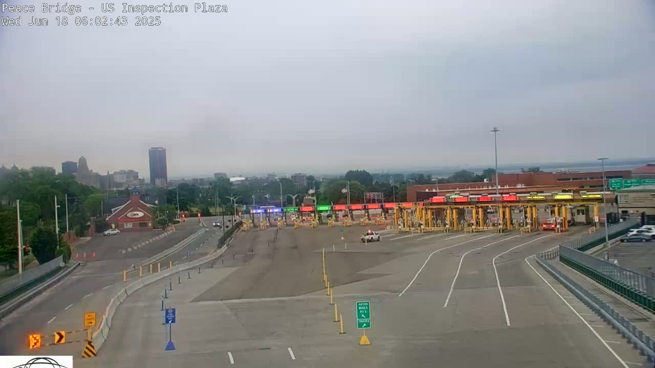 A mostly empty border inspection plaza with a few vehicles is seen on an overcast day, with a city skyline visible in the background.