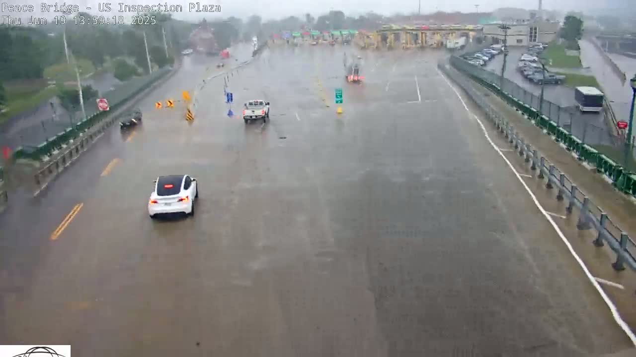 A flooded roadway and parking lot are seen during heavy rain, with several vehicles navigating the water.